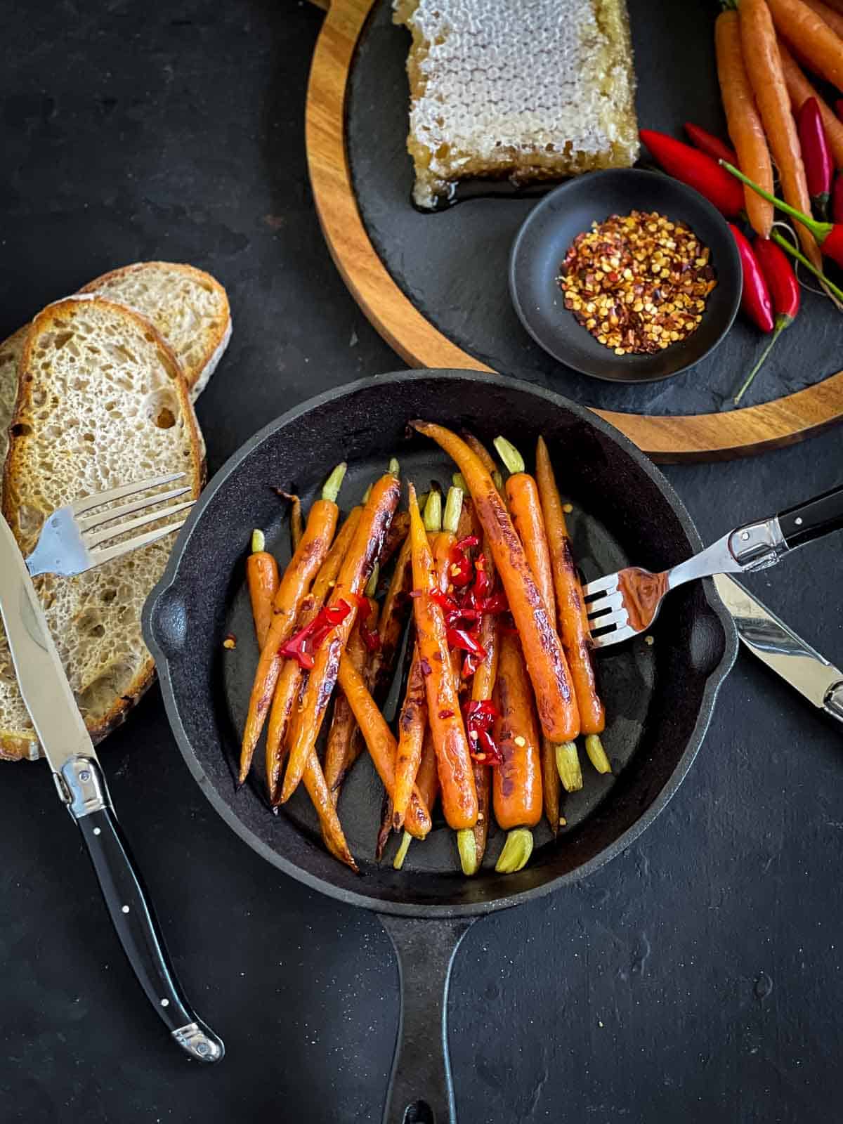 Sous Vide Carrots with Hot Honey in a skillet with some sliced bread, black knife and fork and recipe ingredients to the side.