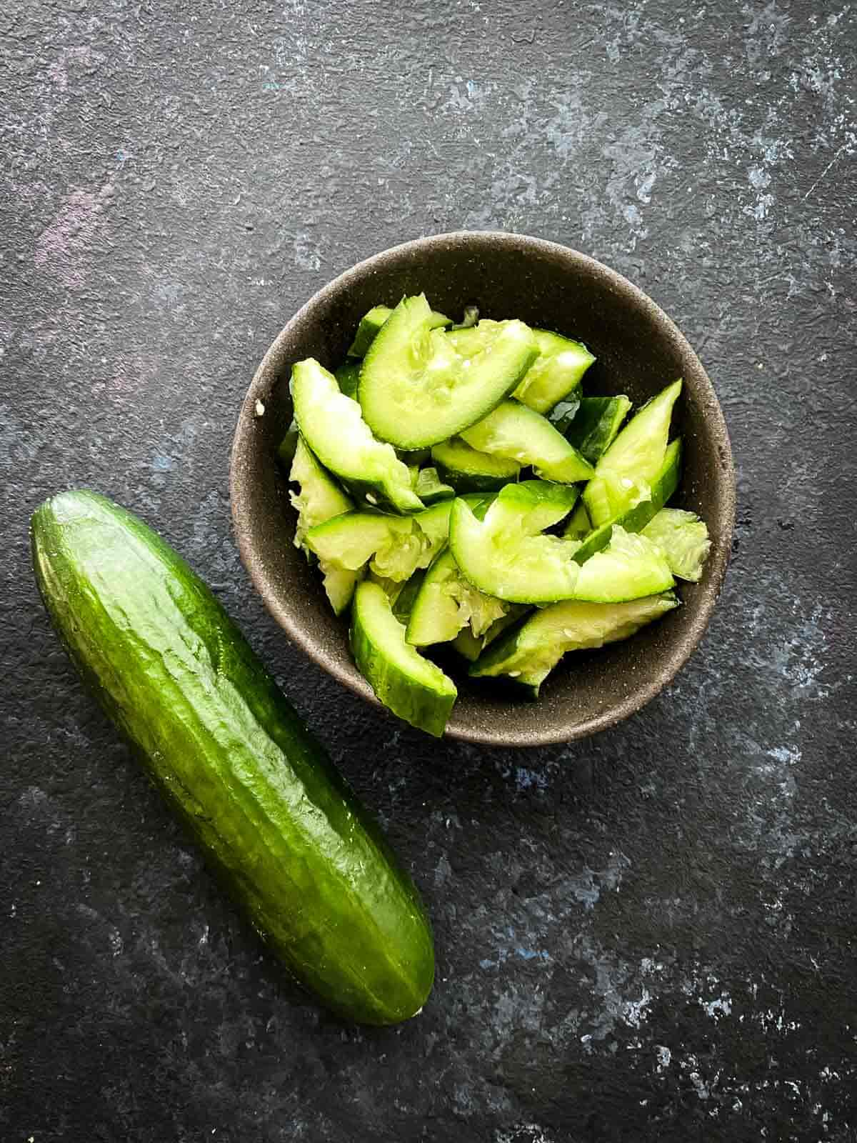 Smashed cucumber in a small brown bowl next to a whole cucumber.