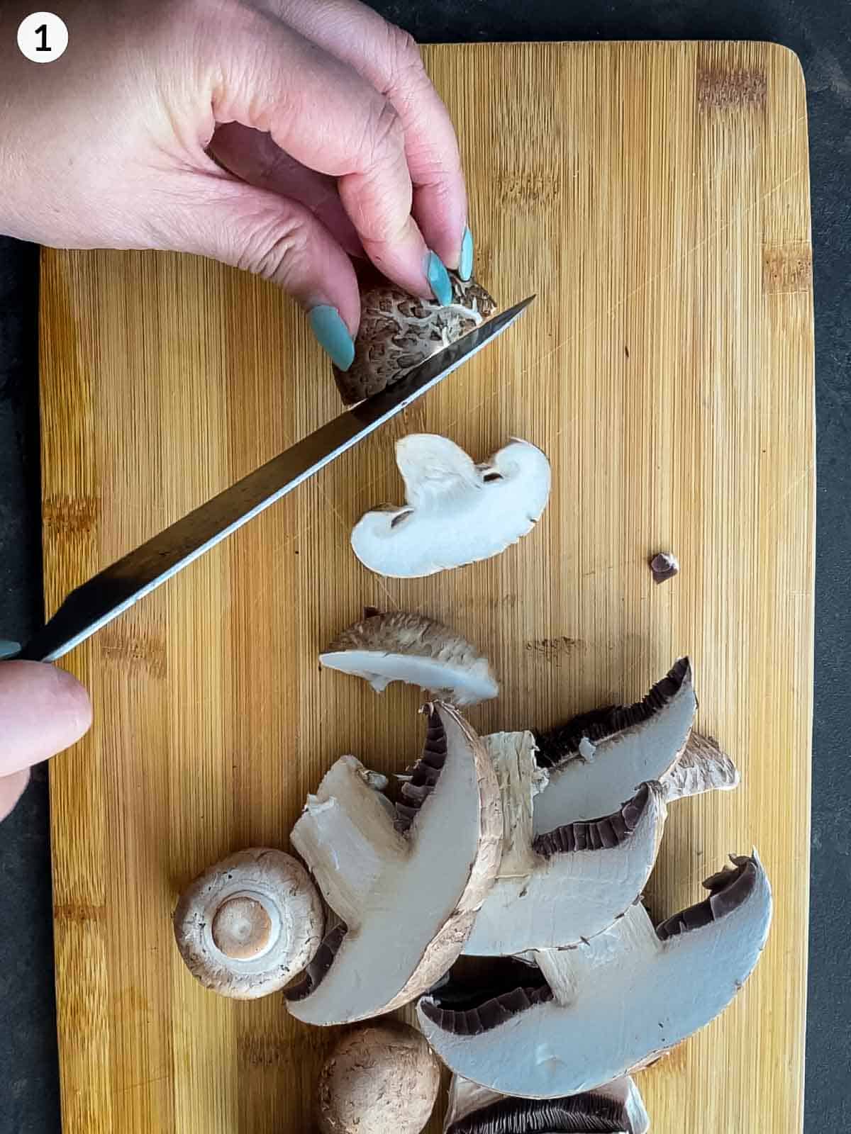 Pair of hands showing how to slice mushrooms with a knife on a wooden chopping board