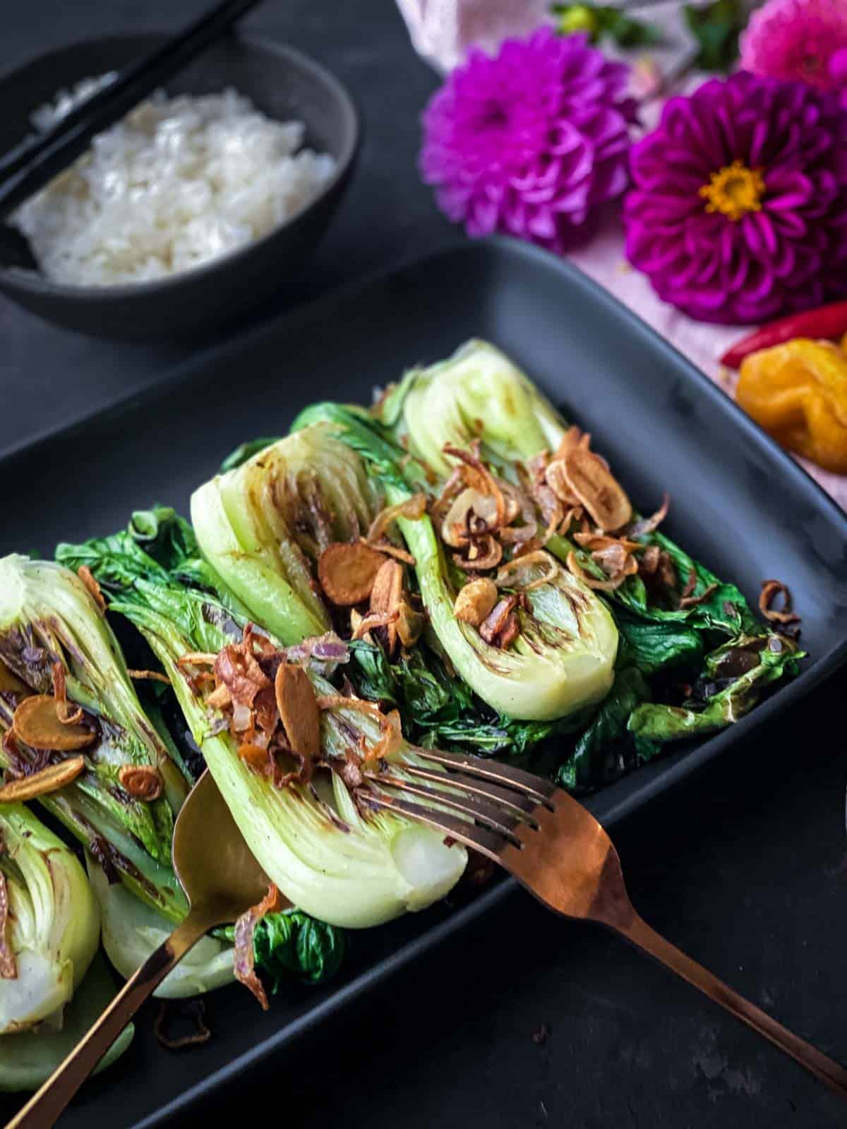 Grilled Bok Choy with Crispy Garlic on a black oblong platter with gold cutlery and pink flowers for decoration