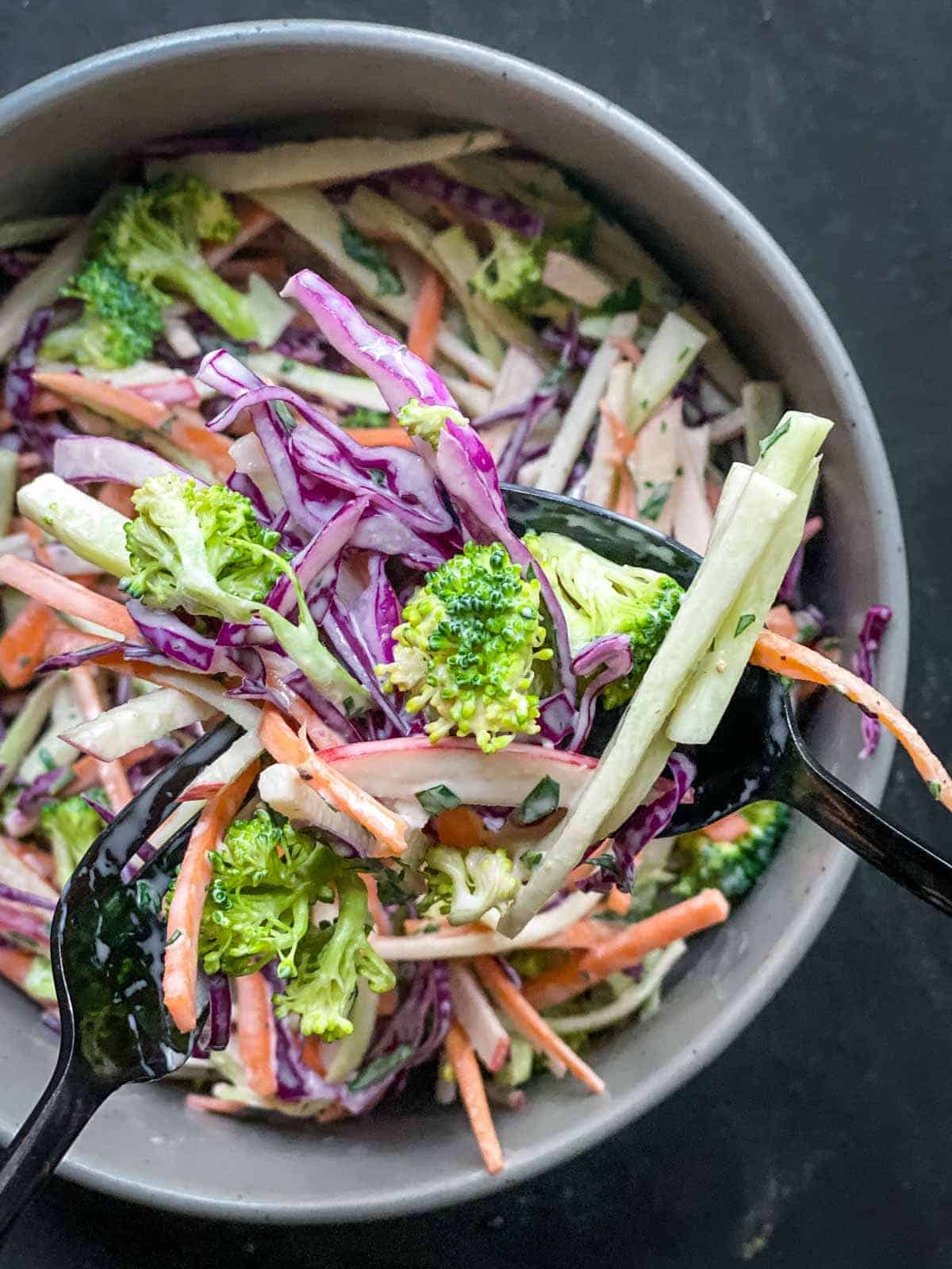 broccoli slaw salad in a grey round bowl with two spoons tossing together