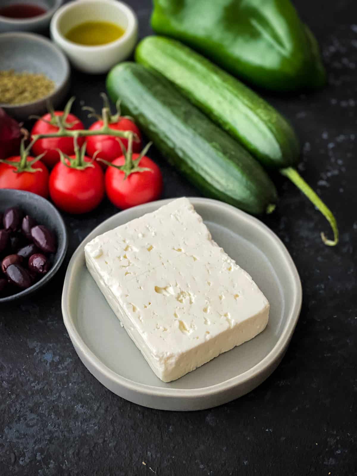 a block of feta cheese on a small grey plate with tomatoes, cucumber, and olives surrounding the plate.