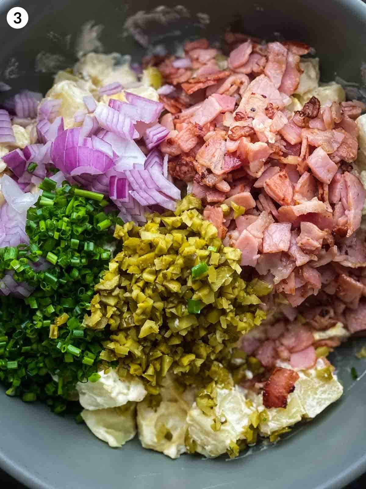 a mixing bowl of diced onions, bacon pieces, pickles, potatoes and chives