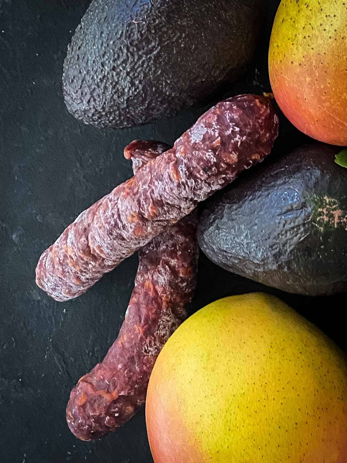 two dry cured chorizo sausages with two avocados and two mangos surrounding them on a black background