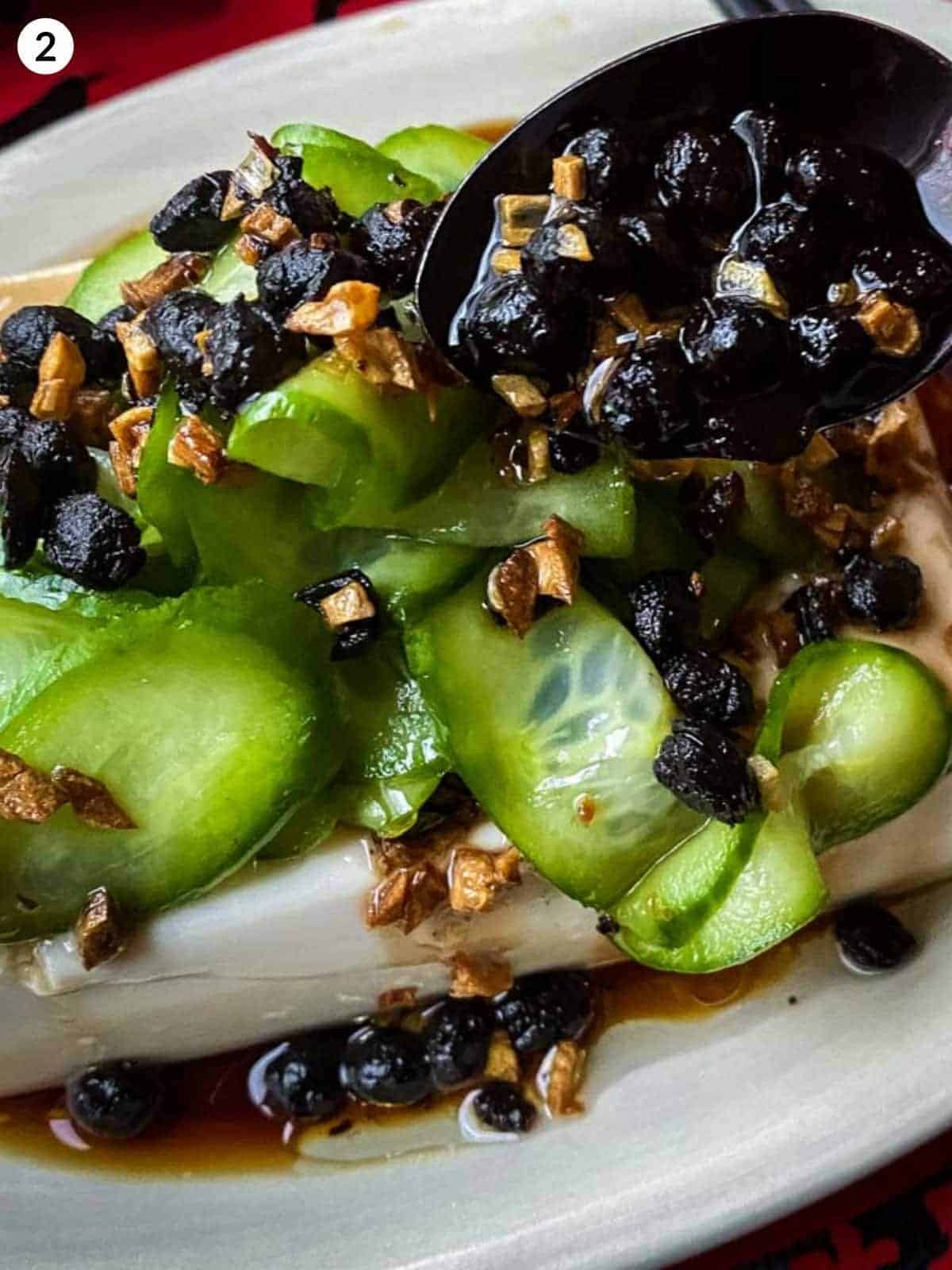Adding fried black beans on top of silken tofu, fried garlic and pickled cucumbers
