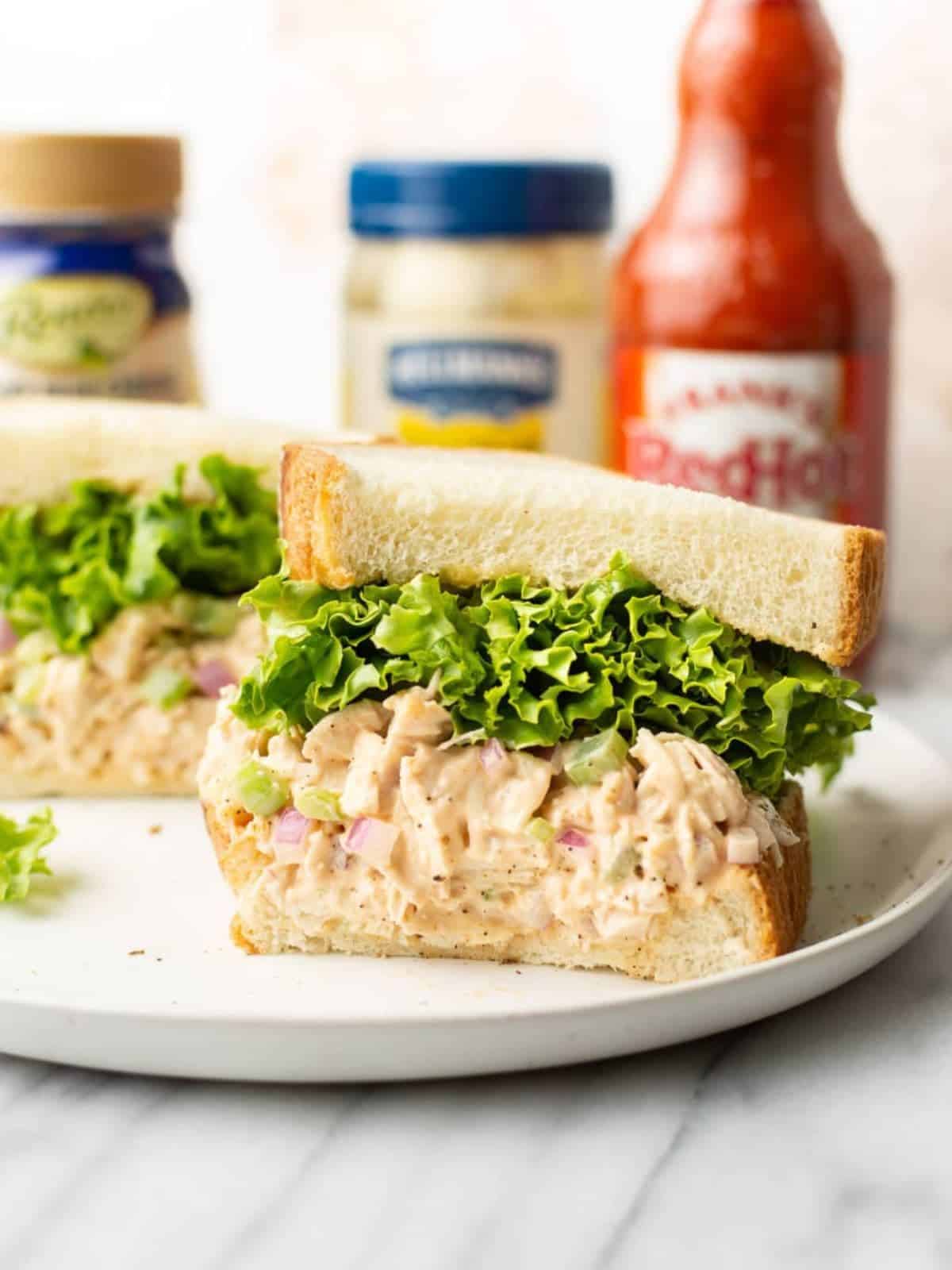 Buffalo chicken salad sandwich on a plate with mayo and red hot sauce in the backgorund