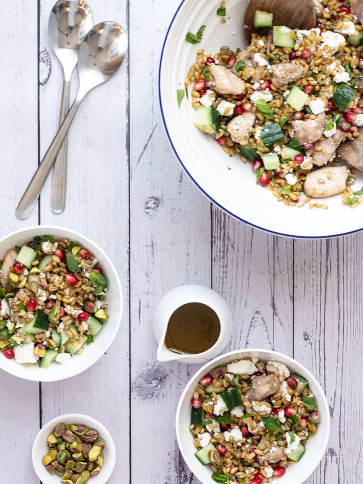 3 serves of chicken and freekeh salad on a white washed wooden table with 2 silver spoons on the side