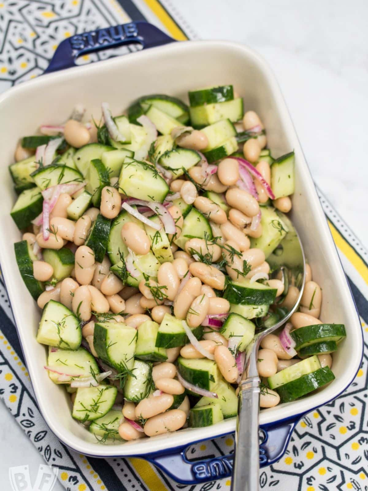 cucumber and cannellini bean salad in a ceramic baking dish on blue and yellow table runner