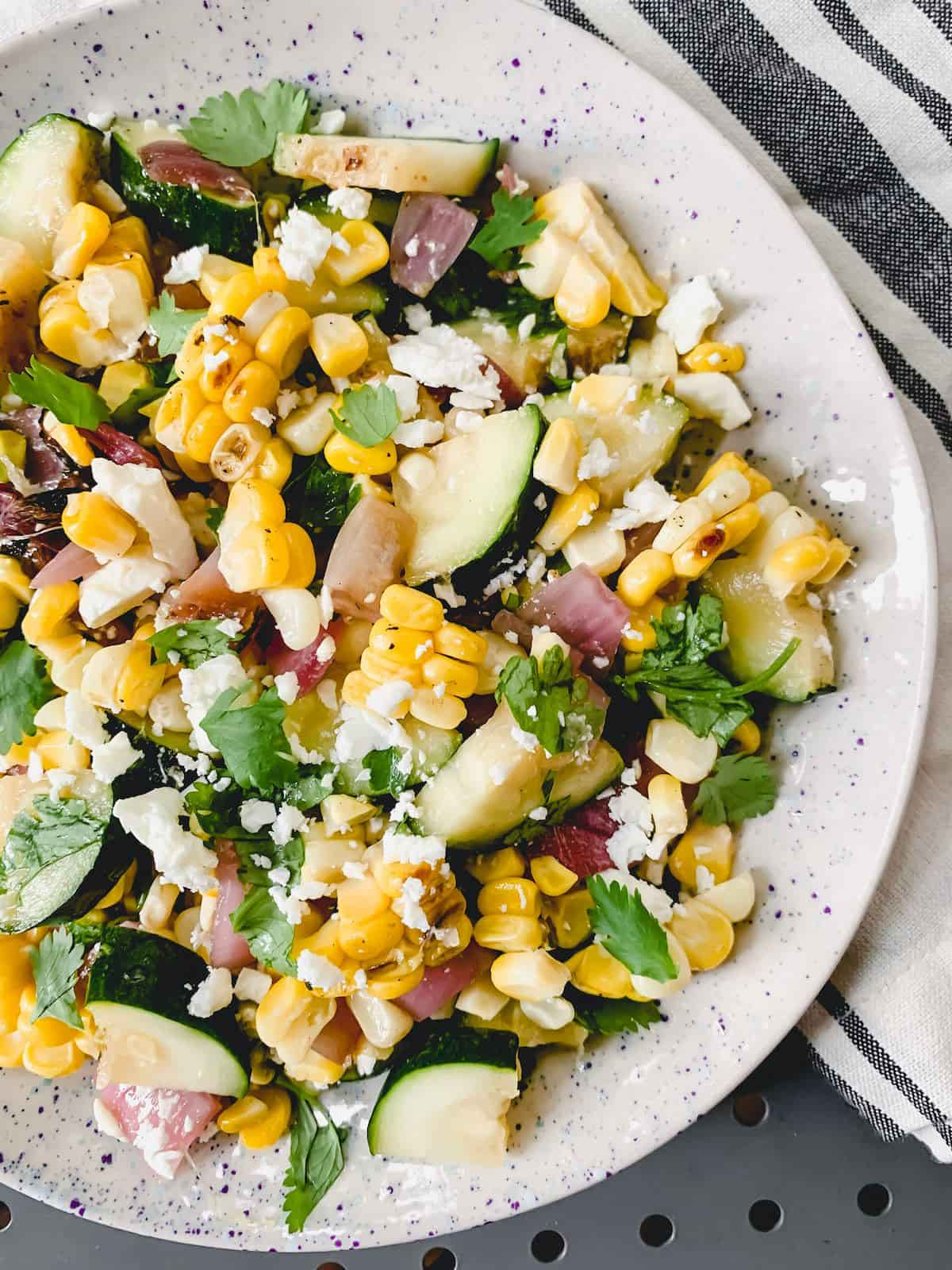 Grilled corn and zucchini salad on a white plate with purple speckles on a blue and white striped napkin