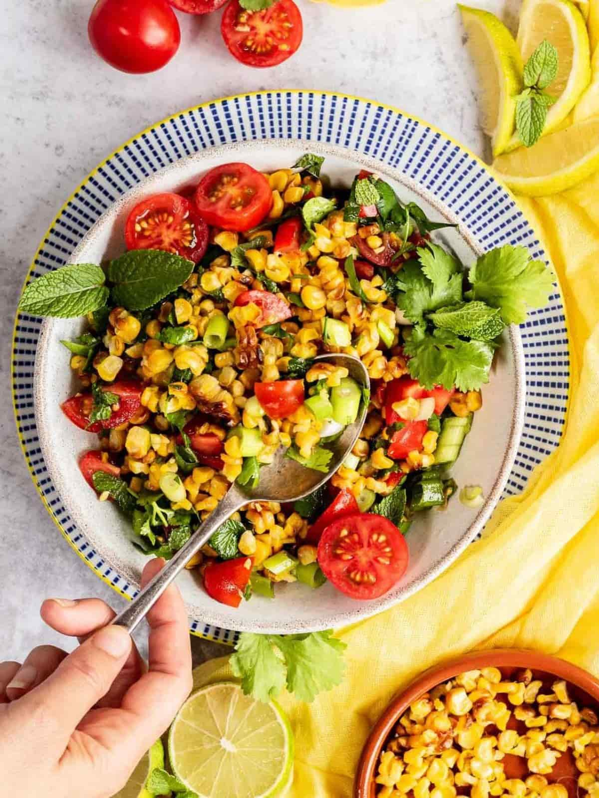 Hand lifting a serve of roasted corn salad with a spoon from a white bowl set on a blue and white pattern plate