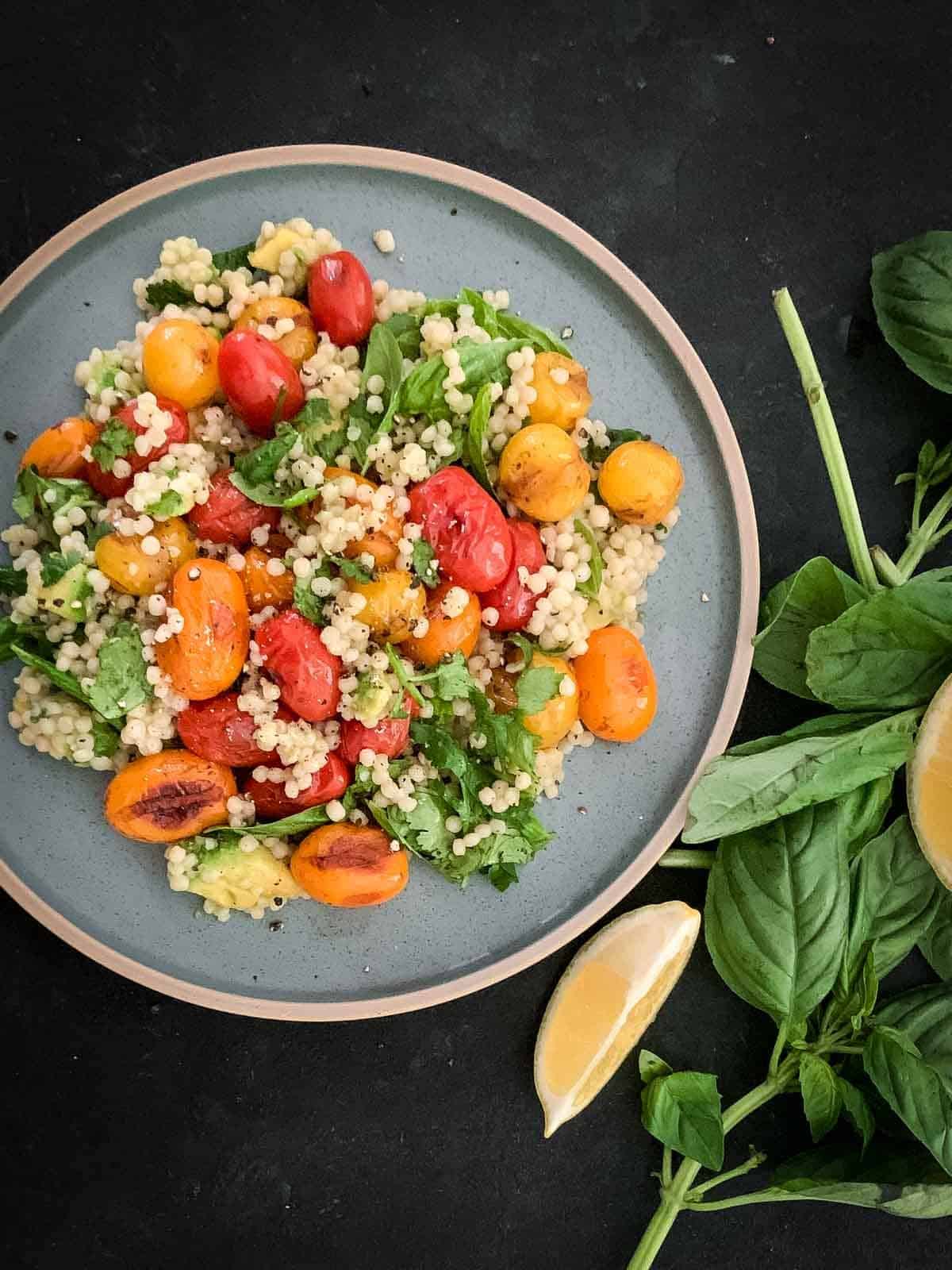 Vegan Couscous Salad with Blistered Tomatoes on a blue plate served with fresh basil and lemon on the side
