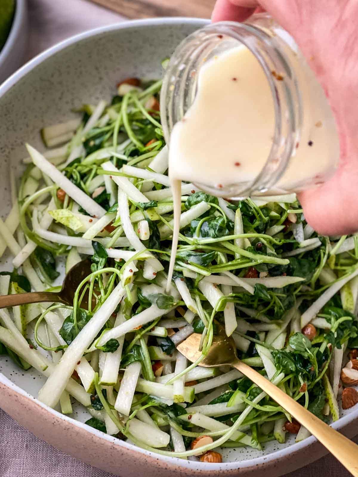 Pouring creamy mustard dressing from a glass jar onto Watercress Salad with Apple and Pear in a grey bowl