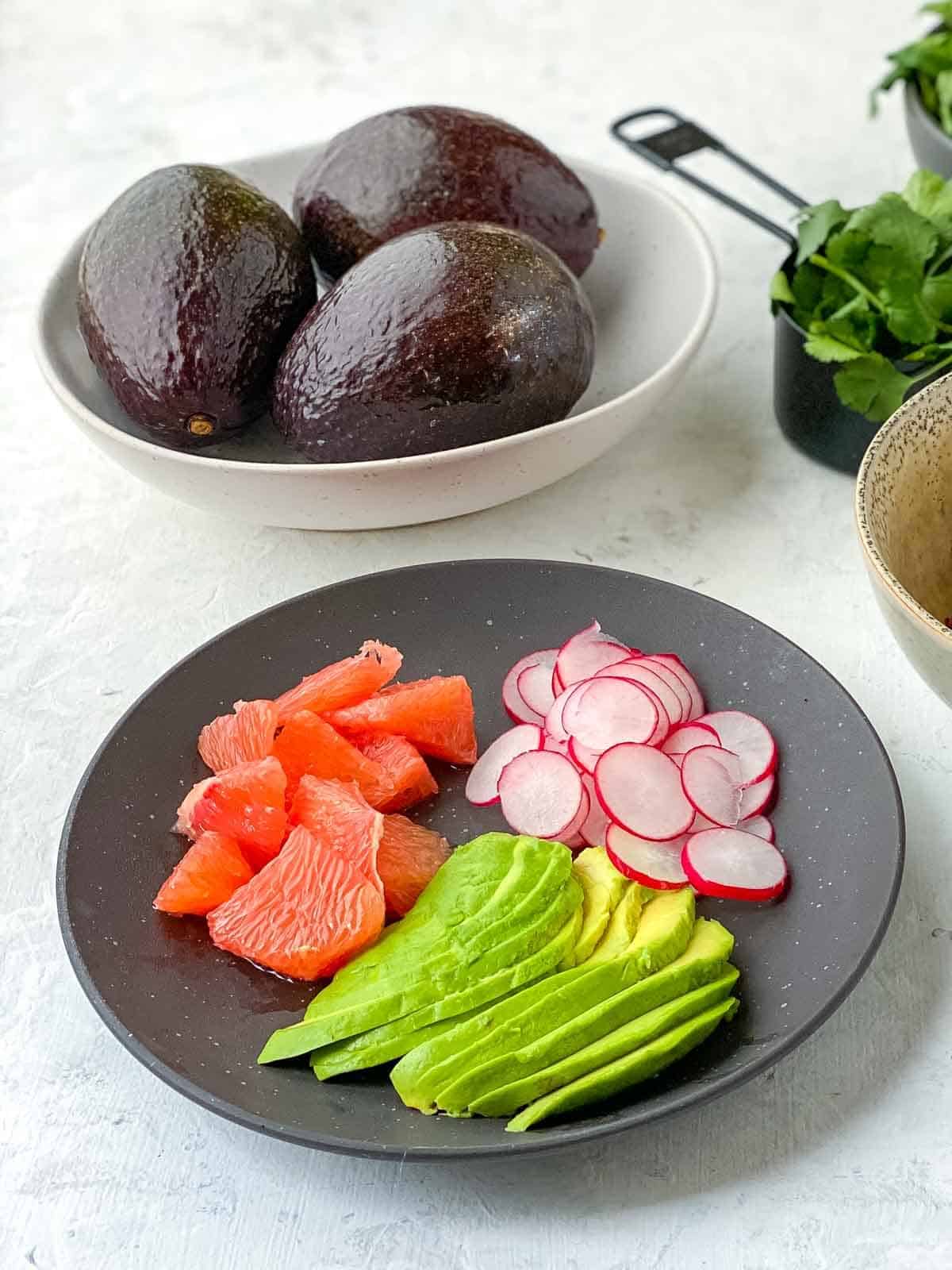 sliced avocado, radish and ruby grapefruit on a black plate with whole avocados in the background