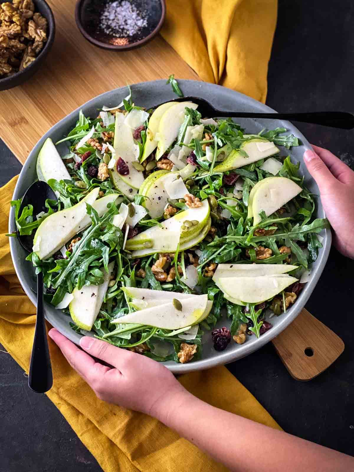 pear arugula salad on large grey plate next to a bowl of candied walnuts and arugula