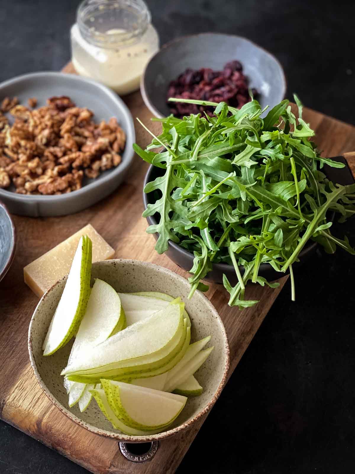 bowl of sliced pear next to bowl of arugula with other salad ingredients in background on a chopping board