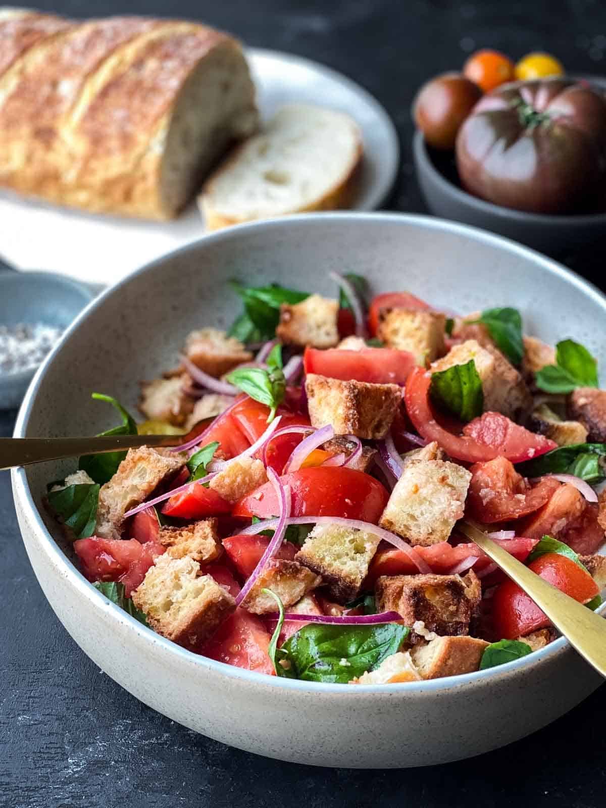 Panzanella salad in a white salad bowl with gold cutlery with sliced bread loaf in the background