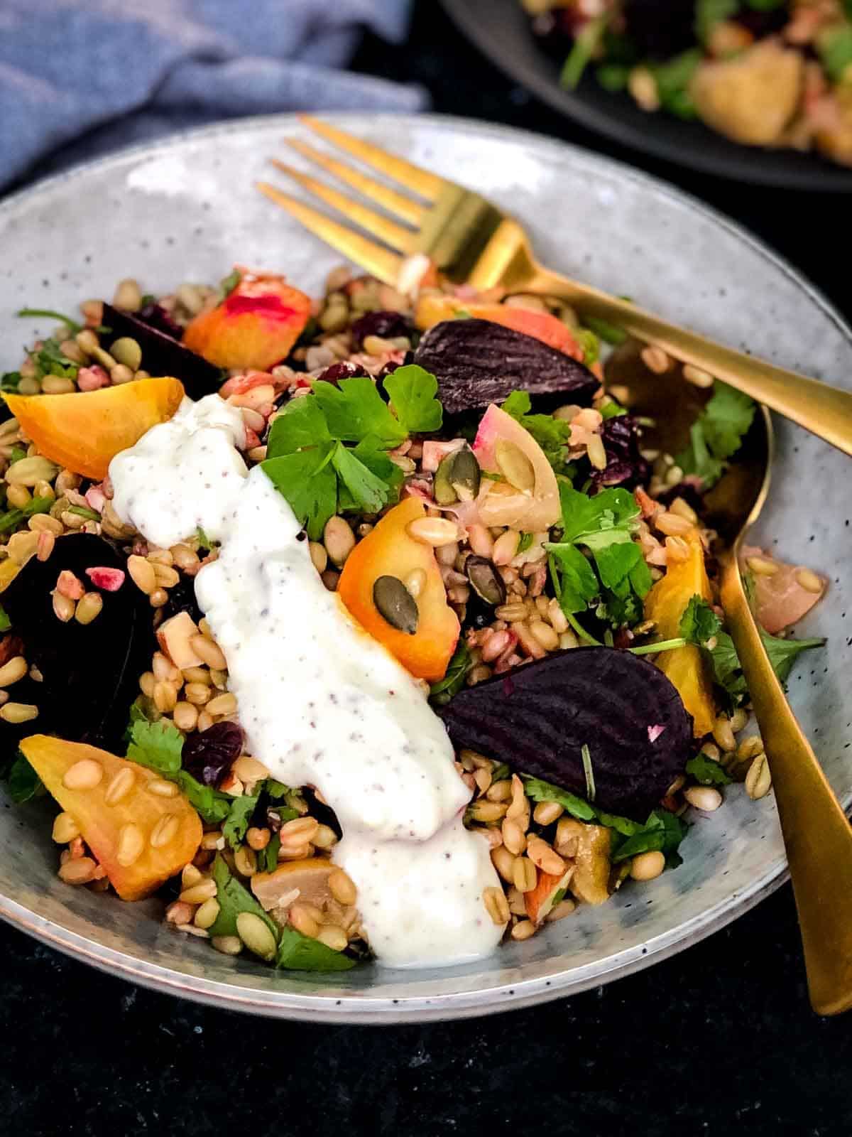 a bowl of freekeh grain salad with roasted beetroot and spiced sumac dressing drizzled on the side. Using gold fork and spoon to serve.