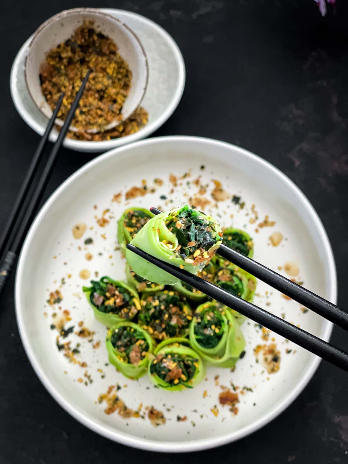 Close up of a Chinese Squash and Spinach Roll held up by a chopstick with furikake in a bowl in the background