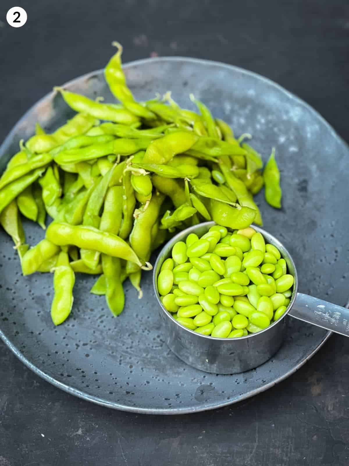 hulled edamame beans in a measuring cup next to edamame in pods on a grey plate