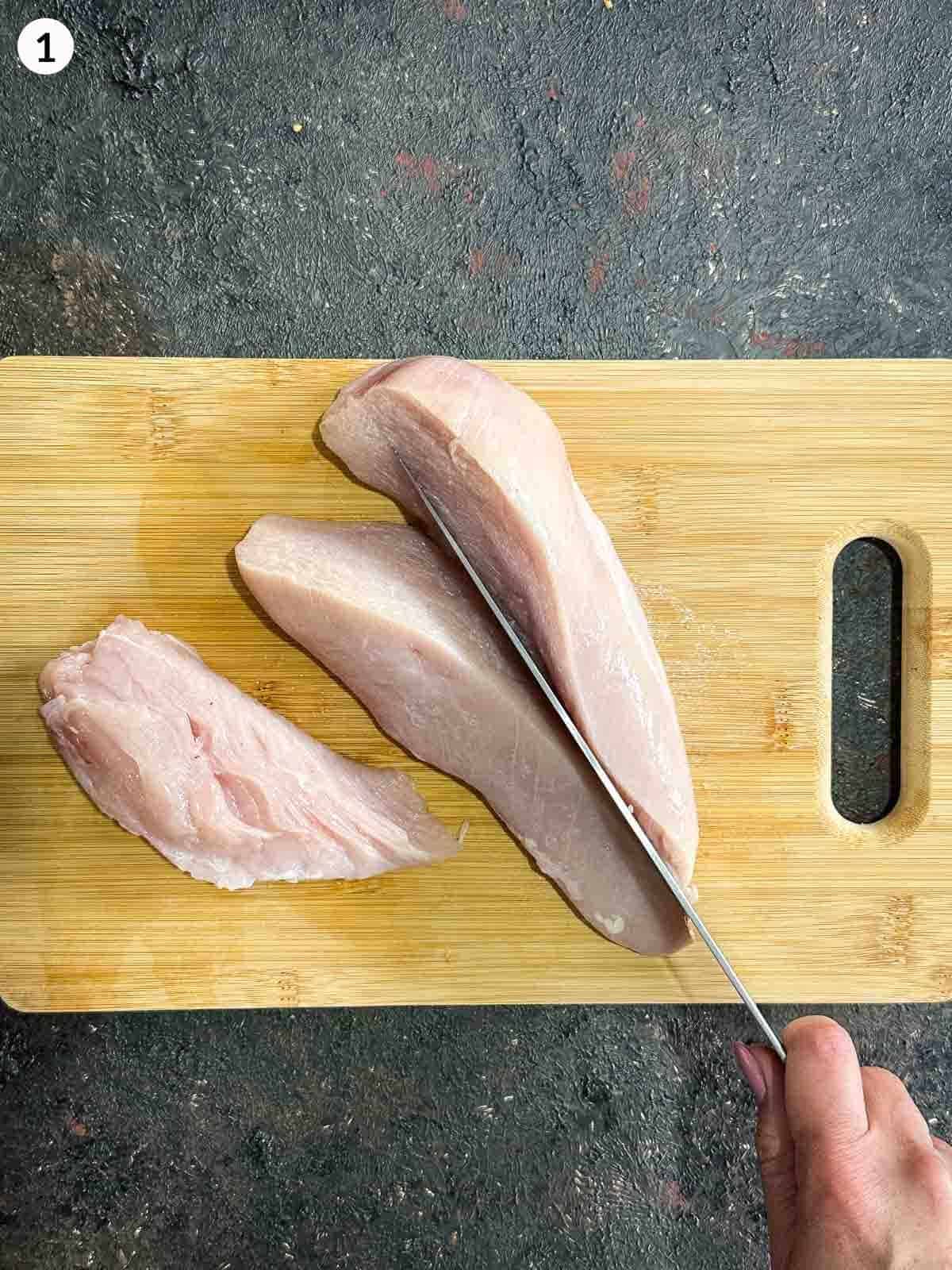 Cutting chicken breast into 3 with a knife on a wooden chopping board