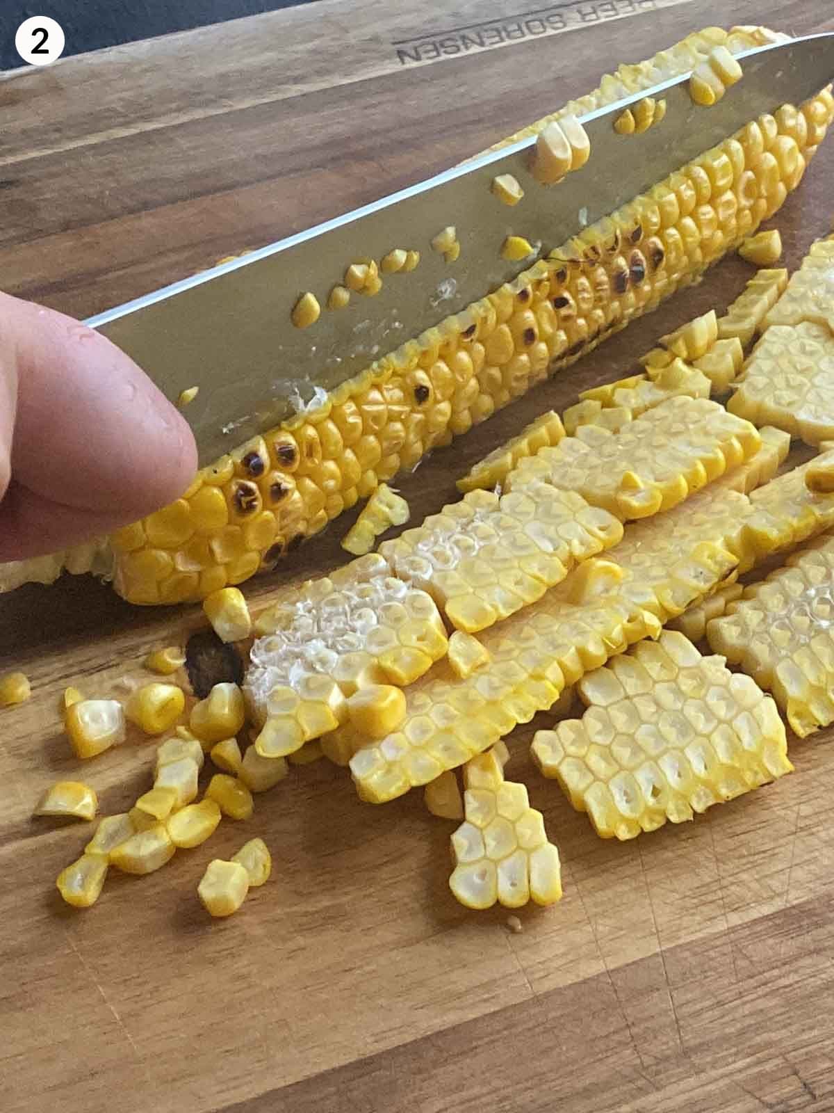 knife cutting corn kernels from cob on a wooden chopping board