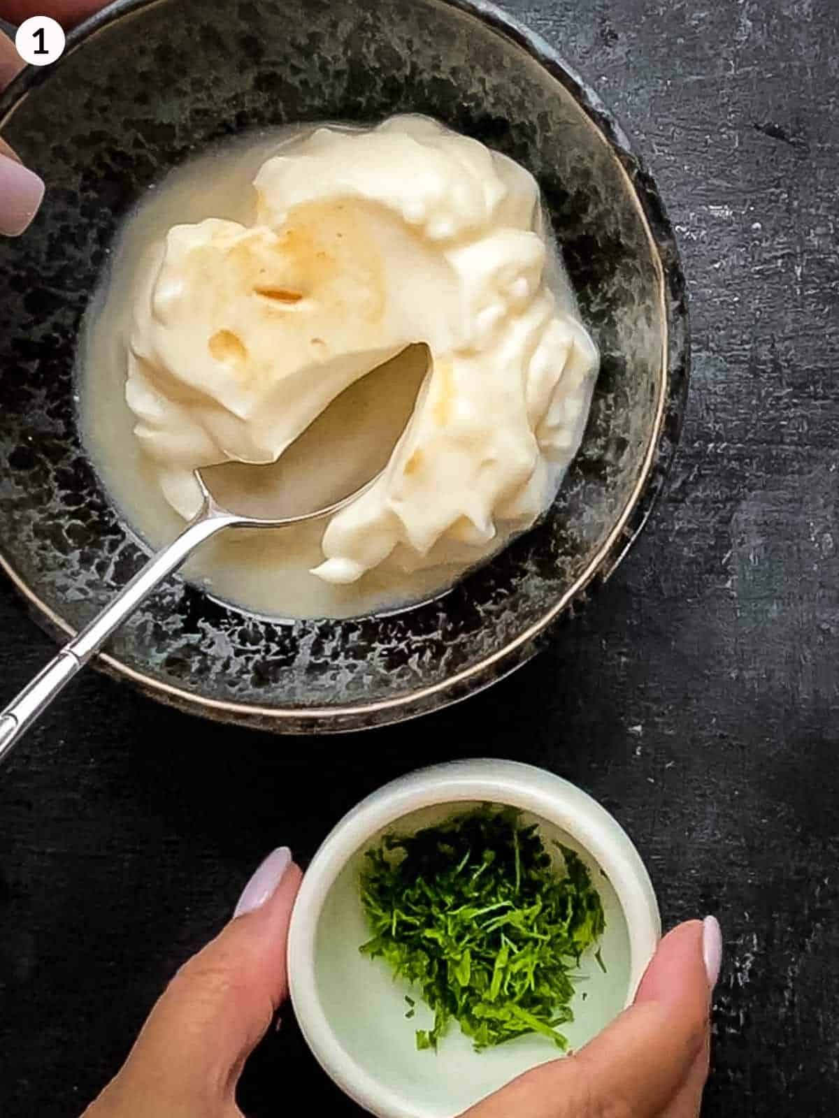 a bowl of mayonnaise, lemon juice and tabasco sauce with a hand holding smaller bowl of chopped dill next to it