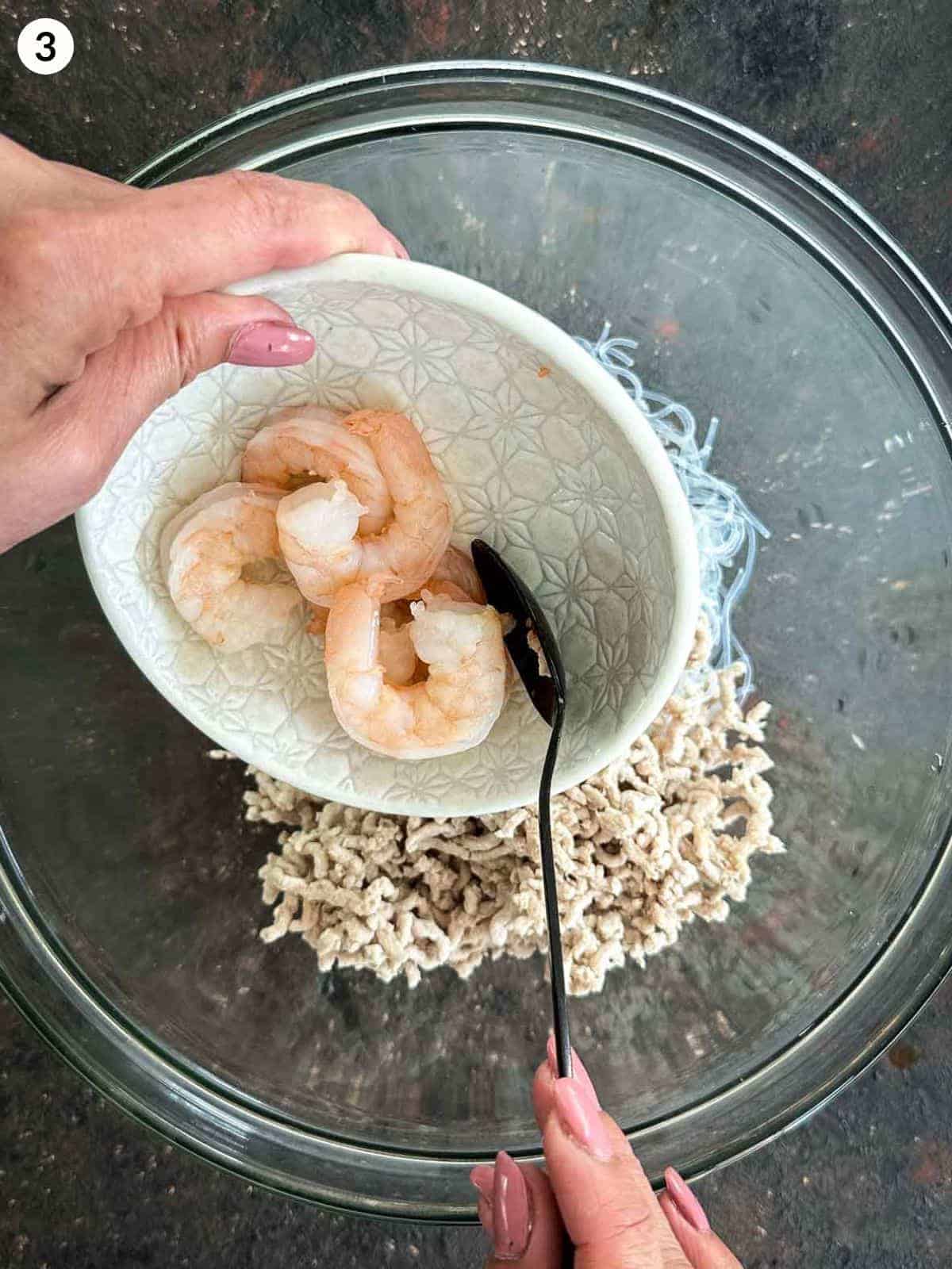 Adding cooked shrimp into a glass bowl of yum woon sen ingredients