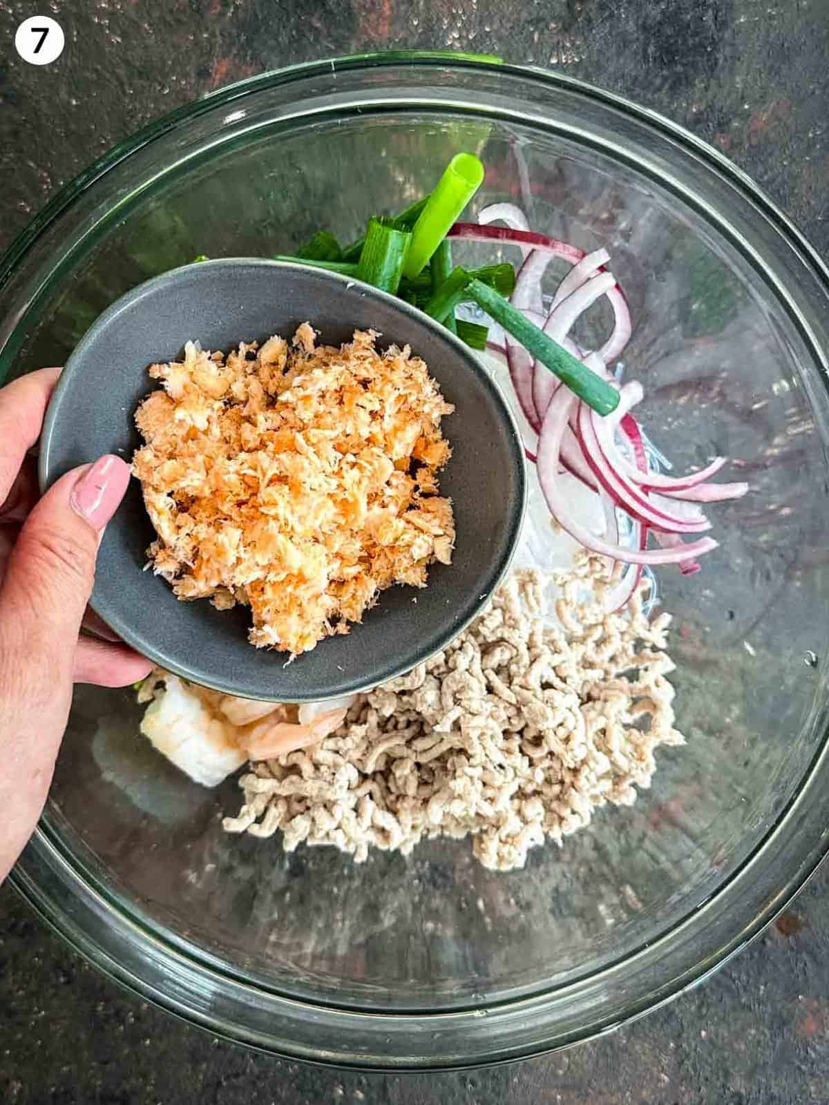 Adding dried shrimp into a glass bowl of yum woon sen ingredients