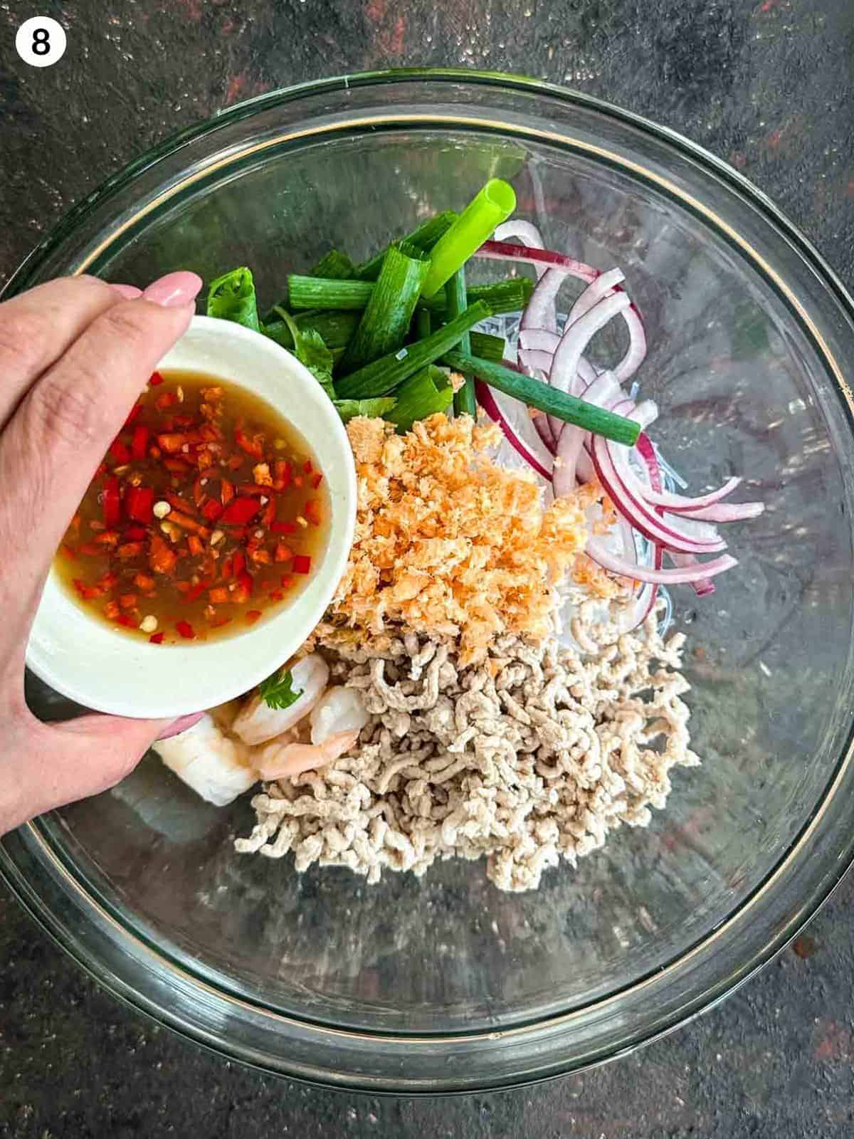 Adding fish sauce dressing into a glass bowl of yum woon sen ingredients