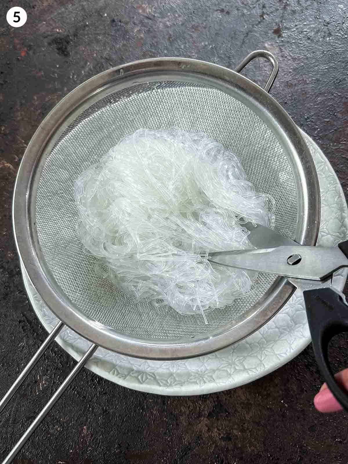 Cutting cooked glass noodles in a sieve over a white bowl with a pair of scissors