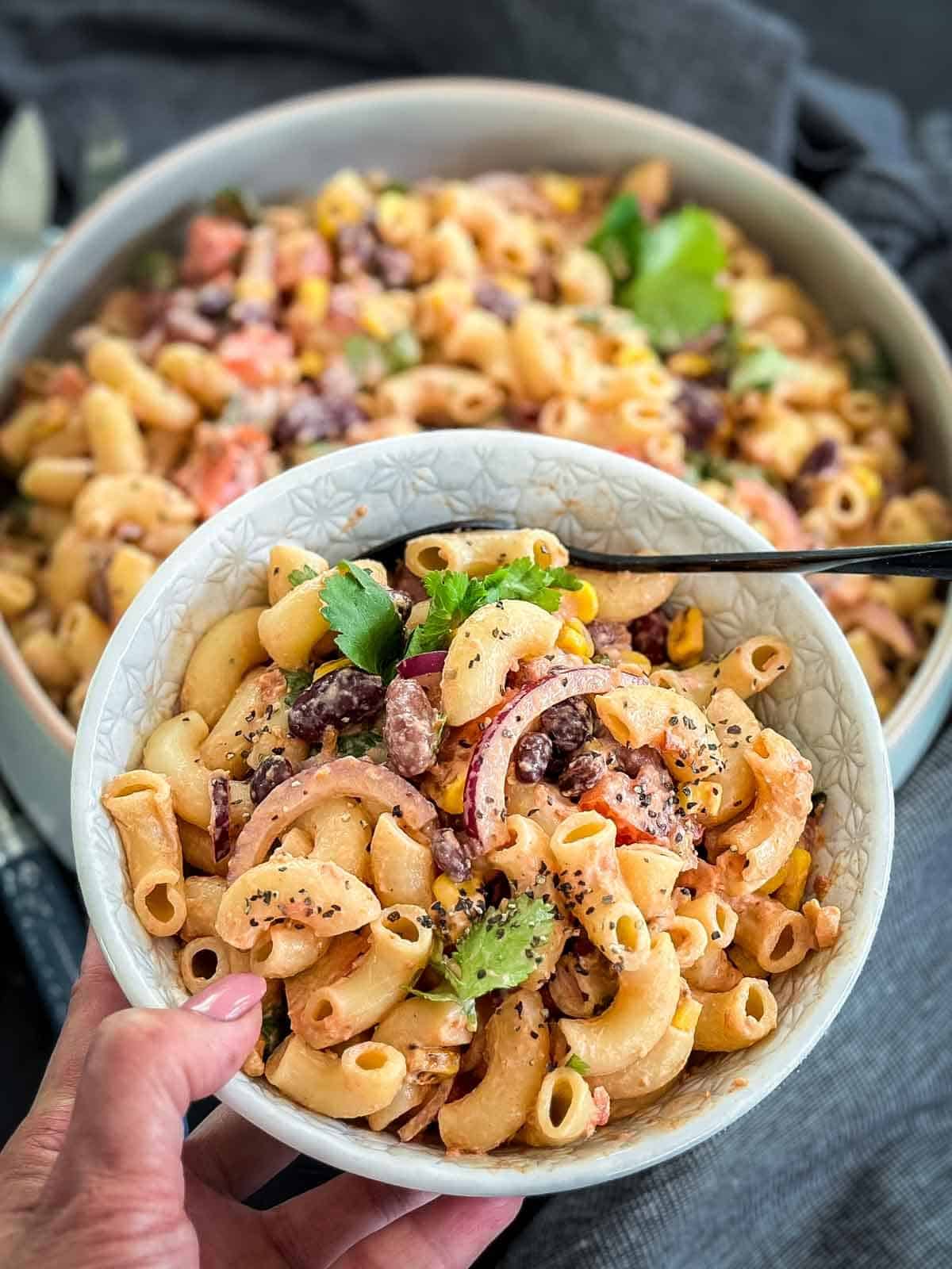 Close up a bowl of Mexican Macaroni Salad [Ensalada de Coditos] hovering over a large bowl of the same salad