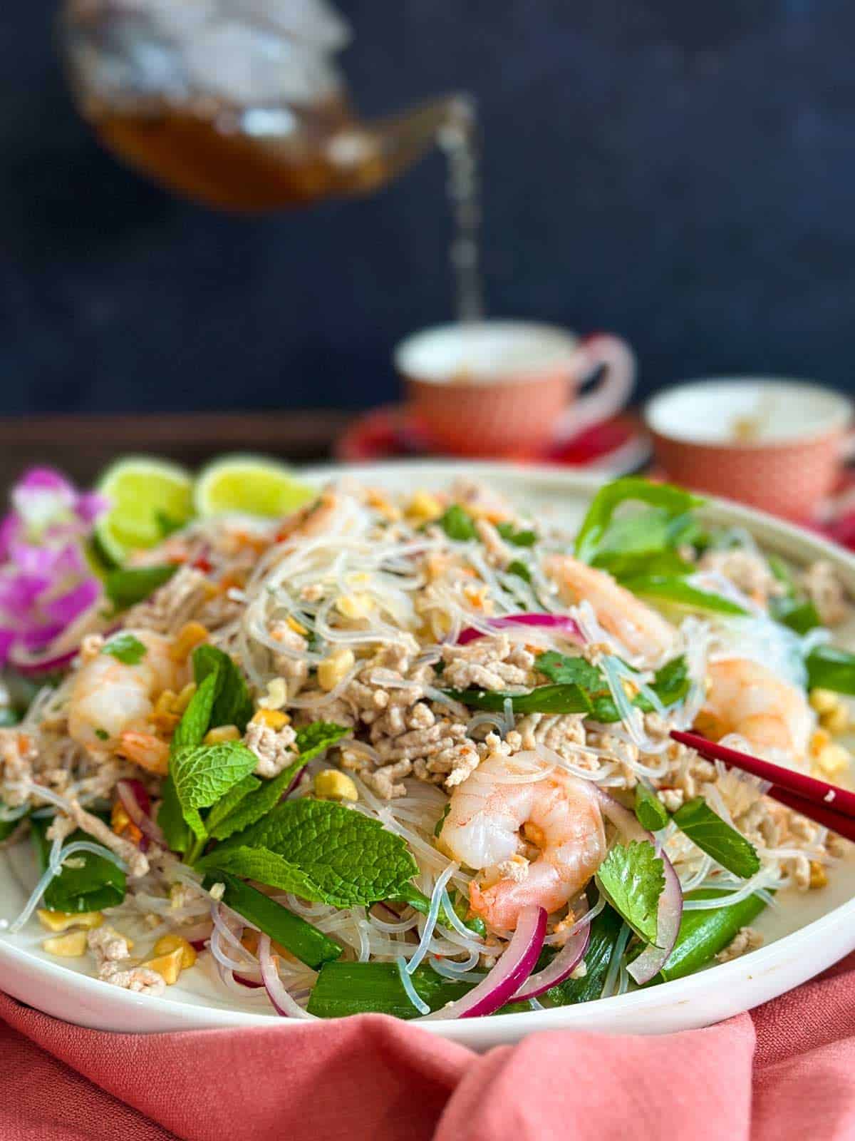 Yum Woon Sen salad on a white plate with pink linen with tea being poured into tea cups in the background