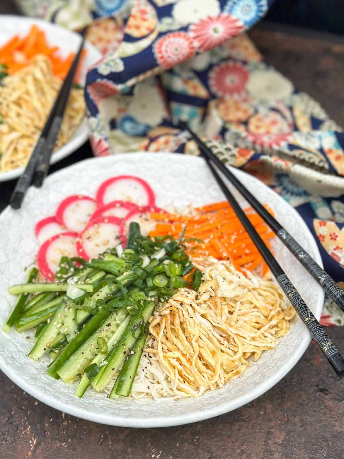 Bowl of somen salad with 2 pairs of black chopsticks and Japanese fabric in the background