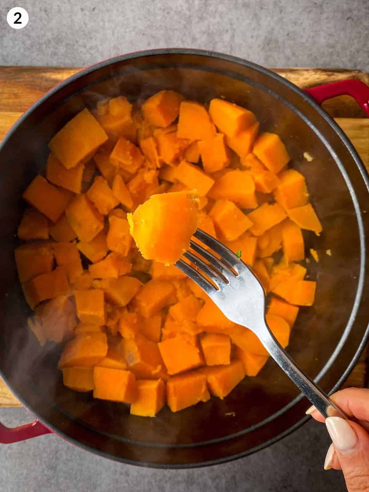 Using a fork to lift up a cube of cooked sweet potato from a dutch oven sitting on a wooden board