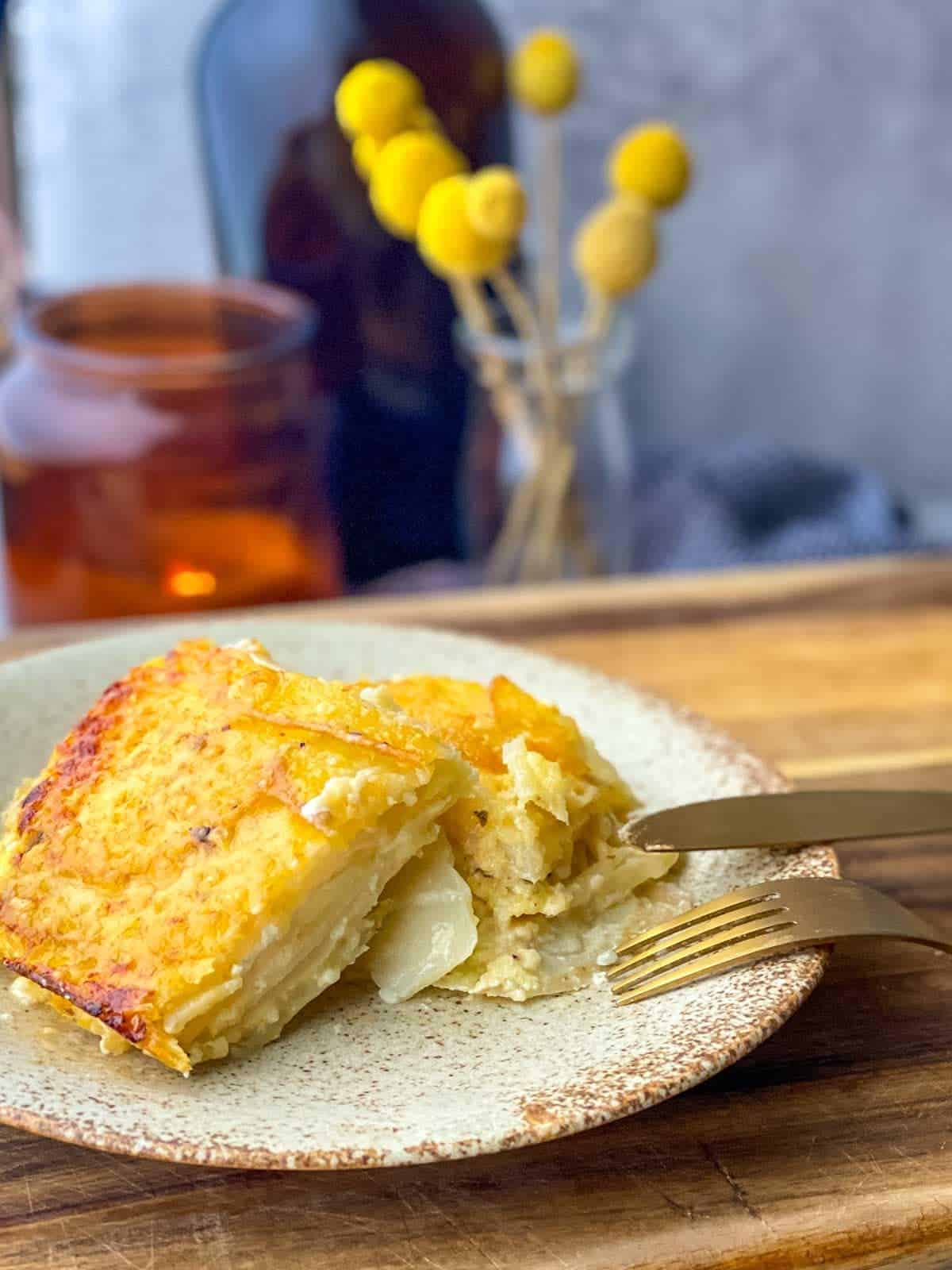 2 square slices of creamy potatoes au gratin on a plate with a knife and fork and dried flowers in the background