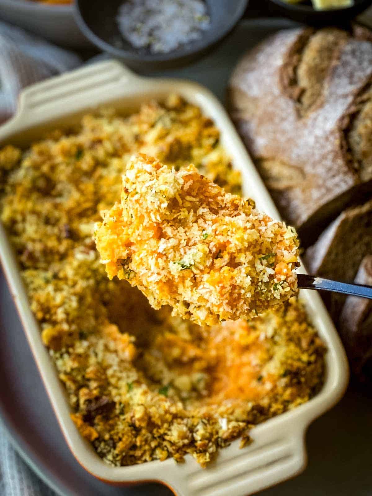 A serving spoon lifting a serve of Savoury Sweet Potato Casserole in a ceramic baking dish, with bread on the side