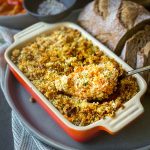 A serving spoon lifting a serve of Savoury Sweet Potato Casserole in a ceramic baking dish, with bread on the side