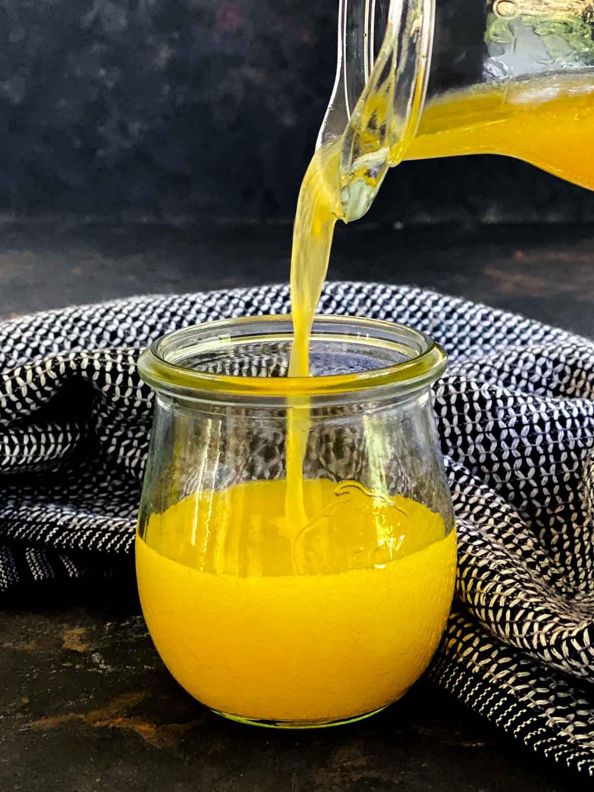 Pouring Apple Cider Vinaigrette with Orange Juice from a jug into a small jar with blue linen in the background