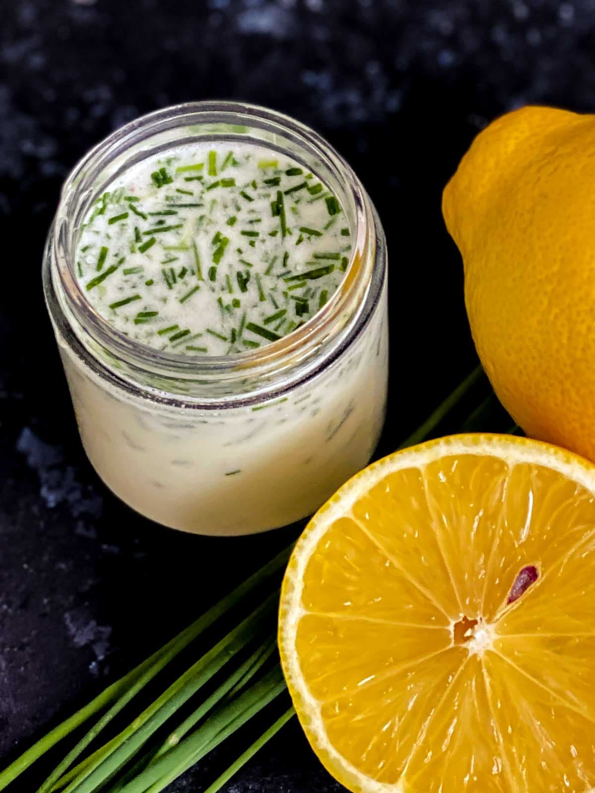 A jar of Creme Fraiche and Chives Dressing with fresh lemons and chives on the side