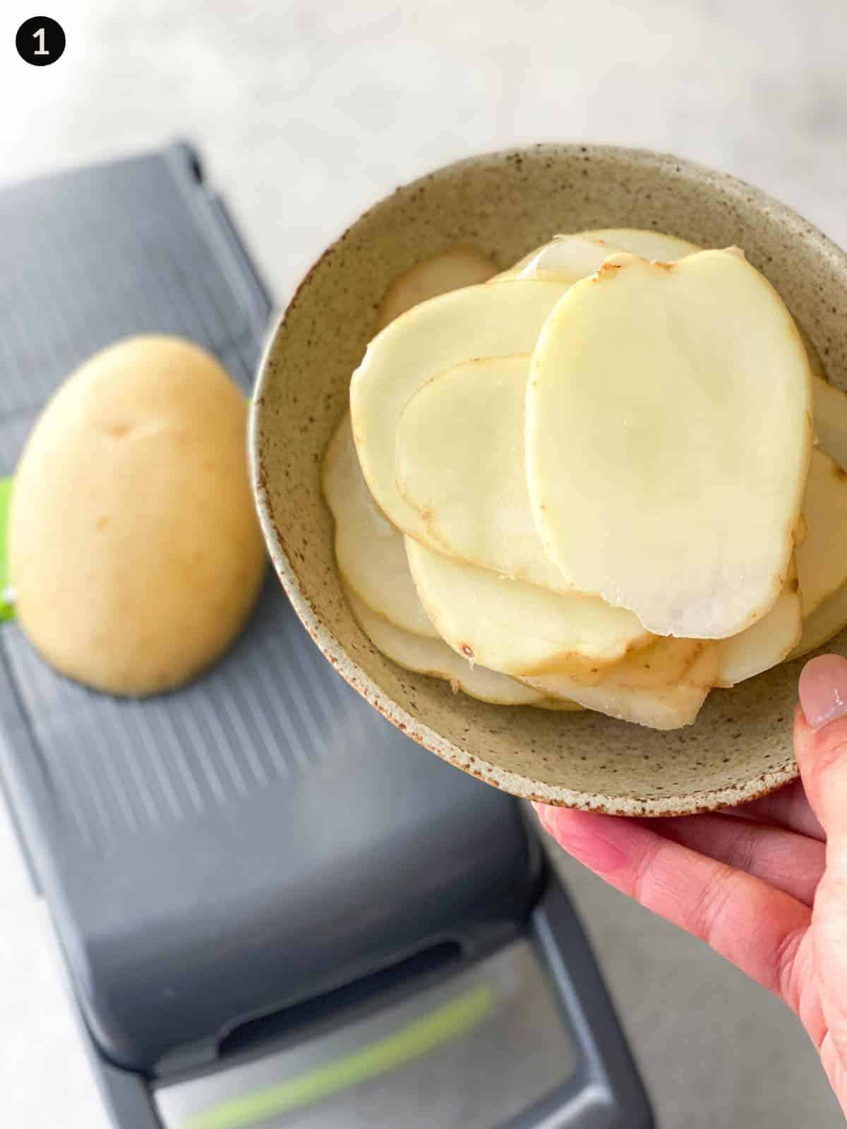 Layers of thinly sliced potatoes in a bowl hovering over a potato on a mandoline