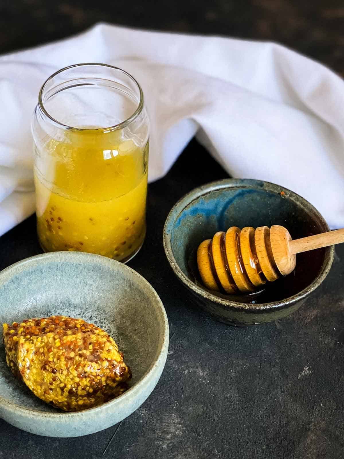 Jar of honey seeded mustard dressing next to a bowl of seeded mustard, bowl of honey with a honey dipper with white linen in the background