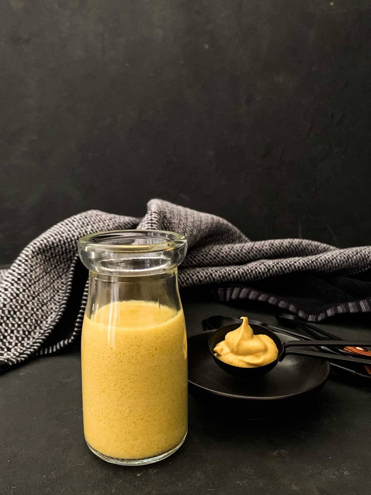 Dijon Mustard Vinaigrette in a clear jar with a measuring spoon of Dijon mustard and blue napery in the background