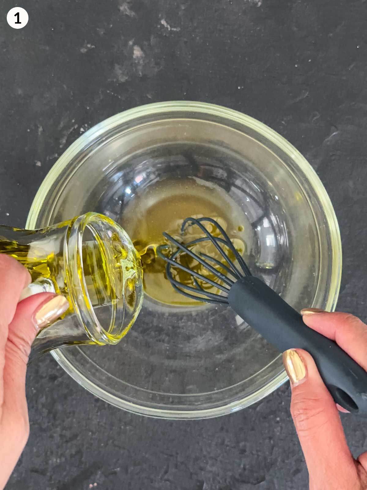 Emulsifying olive oil and apple cider vinegar in a clear mixing bowl with a small whisk