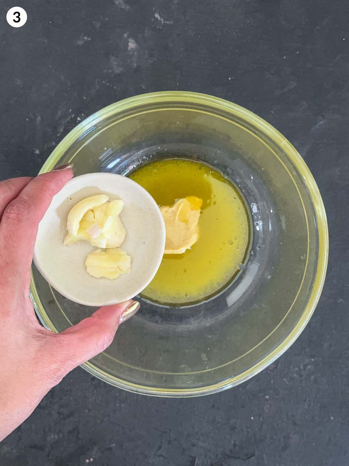 Adding crushed garlic cloves into a clear mixing bowl of salad dressing