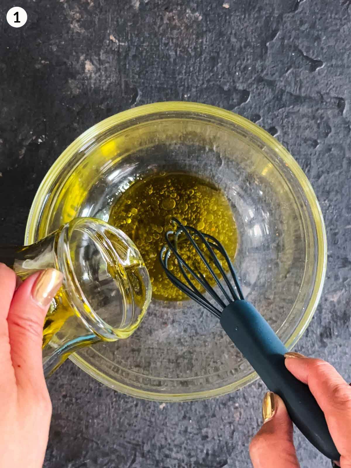 Emulsifying olive oil and apple cider vinegar in a clear mixing bowl with a small whisk