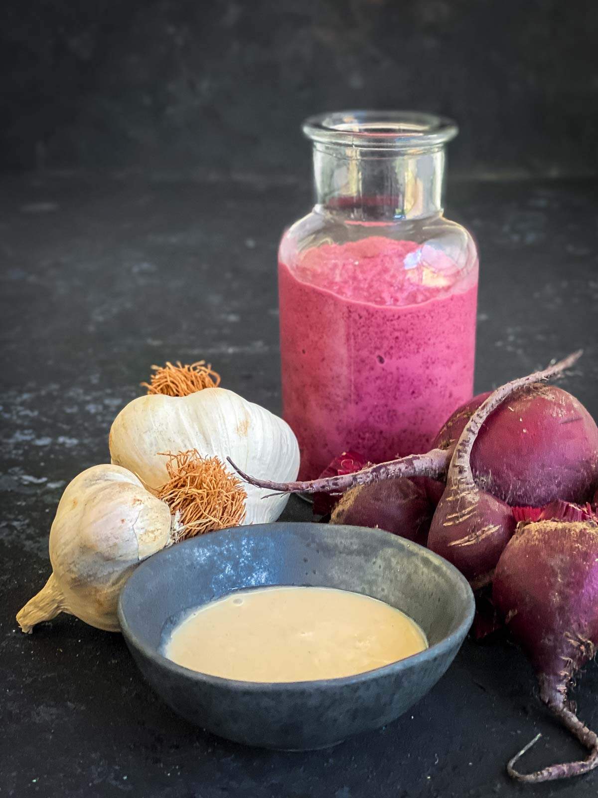 Clear jar of beetroot tahini with whole garlic bulbs, fresh beets and a small bowl of tahini