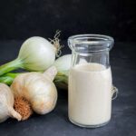 Creamy garlic dressing in a small clear jar with whole garlic bulbs and white onion on the side