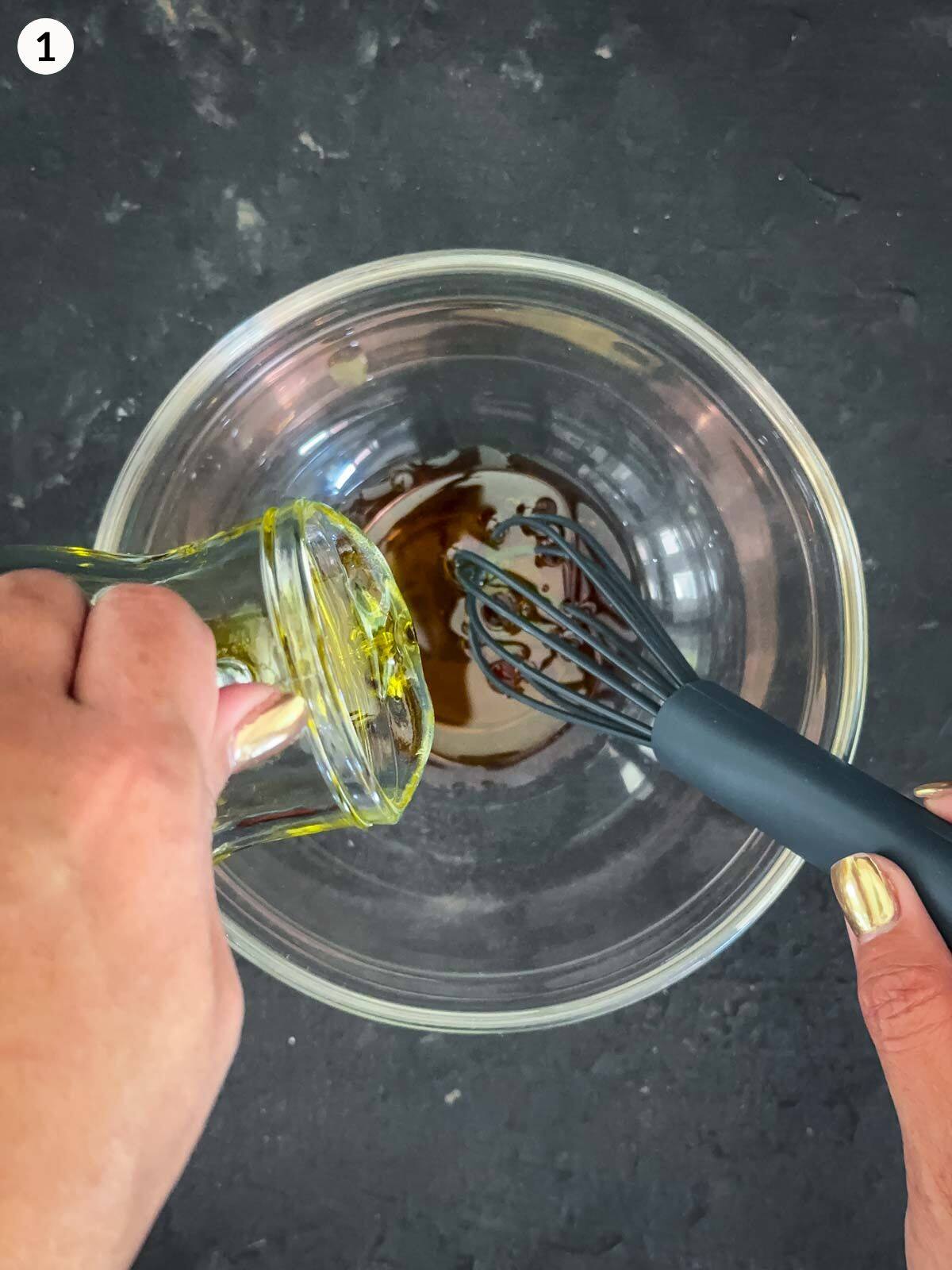 Emulsifying olive oil and red wine vinegar in a clear mixing bowl with a small whisk