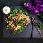 Vibrant oven roasted cauliflower salad with pomegranate seeds and greens, served as a healthy cauliflower side dish with honey Greek yogurt dressing. Salad is served on a black stone board with rose gold cutlery and purple flowers.