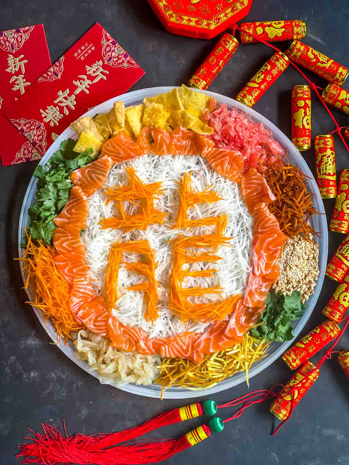 Chinese New Year Yee Sang salad on a large plate with the shredded vegetables spelling out dragon for the year of the Dragon. Surrounded by Chinese red packets and firecracker decorations.